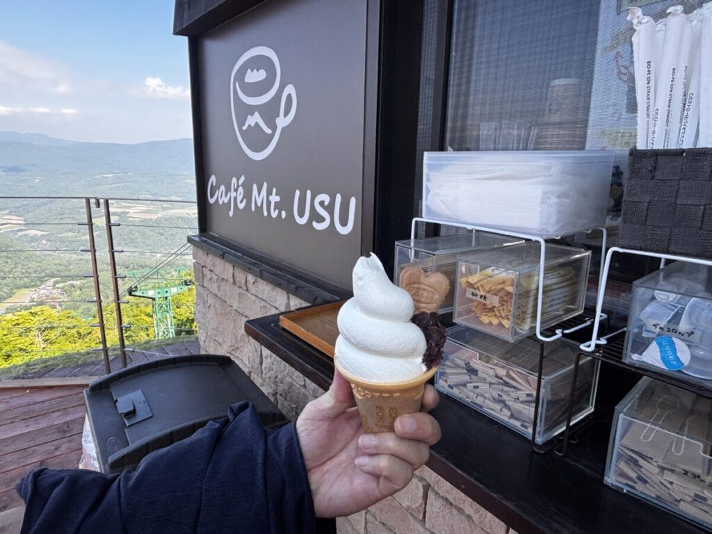 Café Mt.USUで買ったうすもこ牛乳ソフトクリームを山頂の景色を背に手に持つ様子