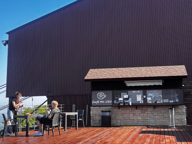 有珠山ロープウェイ山頂駅のCafé Mt.USUの売店とウッドデッキで休憩する人たち