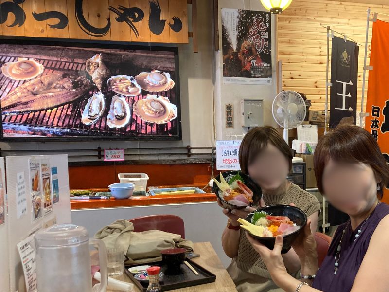 温泉市場の店内で海鮮丼を手にする二人と、炭火焼の写真が飾られた壁