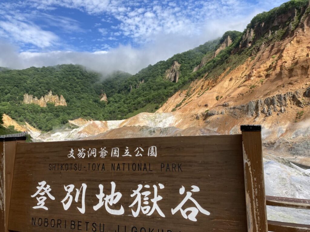 「登別地獄谷」の看板と、うしろに広がる赤茶けた谷と湯けむり